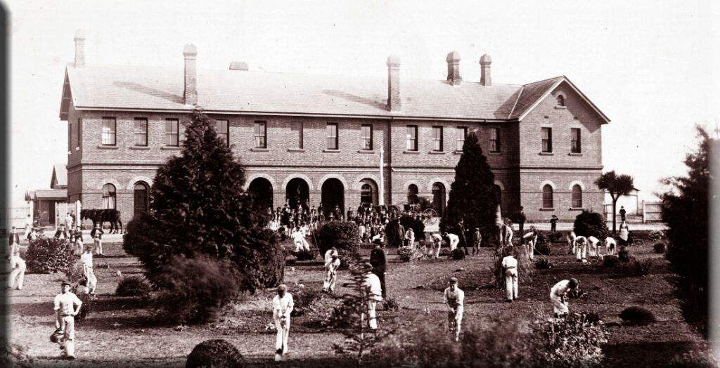 A sepia photograph of the Ballarat Lunatic Asylum. The photo shows the building from the late 1800s, with the grounds filled with patients tending to the large garden in front.
