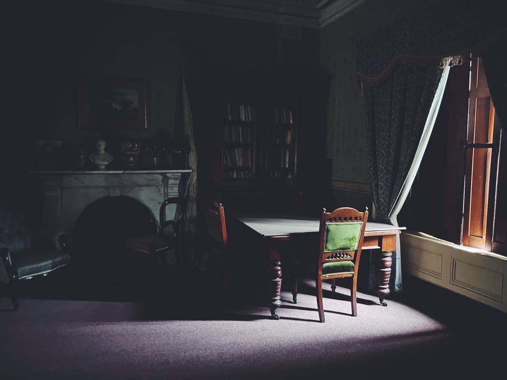 A dark sitting room. A finger of light streams in through an open window, lighting a green upholstered chair at an antique table. 