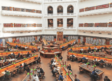 A photo of the inside of the State Library Victoria Dome Room. Long desks stretch our form the centre of the room, people sit at the desks working.