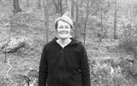 Black and white photo of Louise Falconer,a white woman in a dark coloured top smiling at the camera with a forest in the background