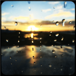 Photograph of the sun rising in a blue sky with low clouds, above a blurry, flat, dark landscape that a shining river is running through. The view is seen from behind a rain spattered window