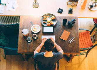 This photo is a birds eye view of a person at their desk writing on their laptop, beside them is a cup of black tea or coffee, a glass of water and a plate of snacks.