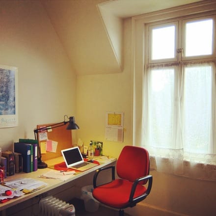 A photo of Studio 5 at Glenfern, with a red chair and a long desk with a laptop, lamp, pin-up board and neat rows of books and files