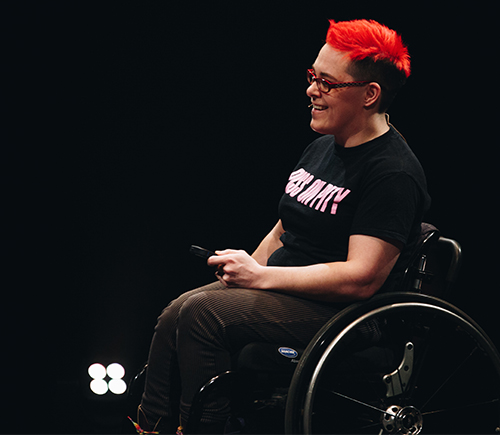 A photo of a woman with bright red hair in a wheelchair. Her T-shirt reads 'Piss on Pity'