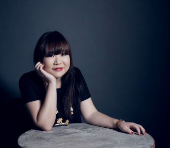  A portrait of Julie Koh, a person with Chinese-Malaysian heritage, pale peach skin and dark hair with a fringe. She is sitting with her chin on her hand.