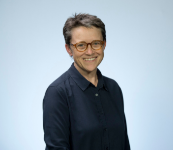 A portrait of Kelly Gardiner, a woman with pale peach skin and short grey hair. She is wearing tortoiseshell glasses and is smiling.