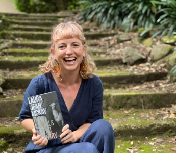 A portrait of Laura Jean McKay, a woman with light peach skin and blonde hair. She is smiling and sitting on stone steps