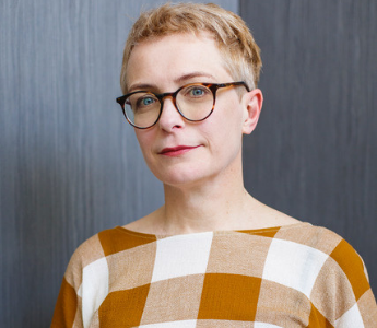 A portrait of Monica Dux, a person with pale peach skin, blue eyes and short hair. She is wearing glasses and looking directly at the camera