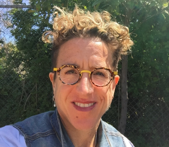A portrait of Nadine Davidoff, a person with medium peach skin with short curly hair. She is wearing glasses and smiling.