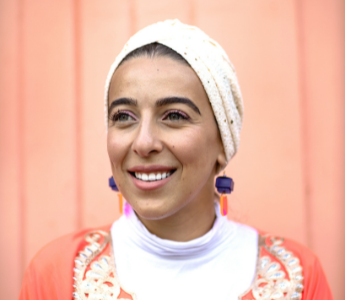 A portrait of Sara Saleh, a person with medium peach skin, wearing a light coloured head scarf and colourful earrings.