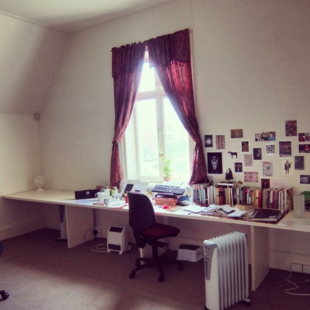 A photo of Studio 4 at Glenfern, with  with a long desk beneath a red-curtained window. The desk is covered in books, tea cups and writing research. The wall is covered in images. 
