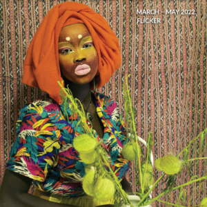 A cover image which is a self portrait of the artist, Atong Atem. She stands staring at the camera. She is wearing an orange scarf around her head, bright yellow and ping makeup, a bright colourful top and a yellow skirt. She is holding a green pot plant. The background is a textured wallpaper. The text reads "The Victorian Writer: March - May 2022. Flicker".