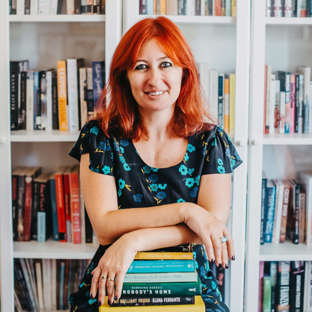 Lee Kofman stands in front of a bookshelf, leaning on a stack of books.