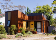 A wood-panelled house sits in a sunny location surrounded by tall trees with lots of greenery.