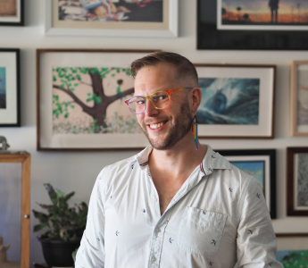 Michael Earp smiles at the camera. They are wearing rainbow glasses and earrings and standing in front of a wall of pictures.